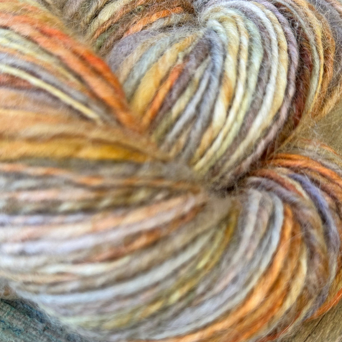A close-up of a twisted skein of variegated yarn resting on a rustic wooden surface. The yarn displays a soft, fuzzy texture with blended stripes of orange, yellow, beige, brown, and muted gray.